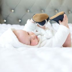 Baby liegt im Bett und trägt dunkelblaue Wildlederpantoffeln.